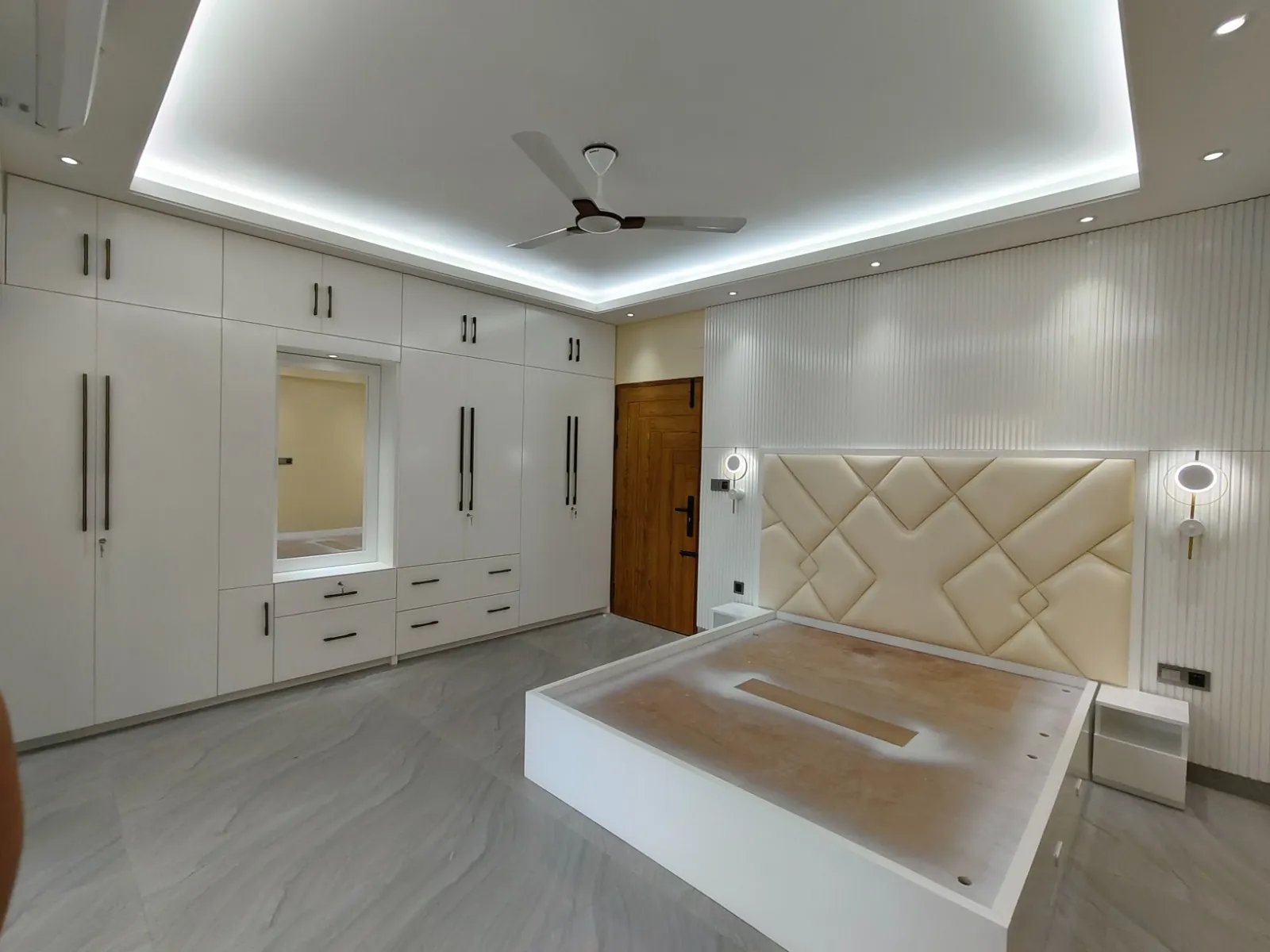 Spacious modern bedroom with white wardrobe, beige upholstered bed with LED wall lights, wooden door, and ceiling fan